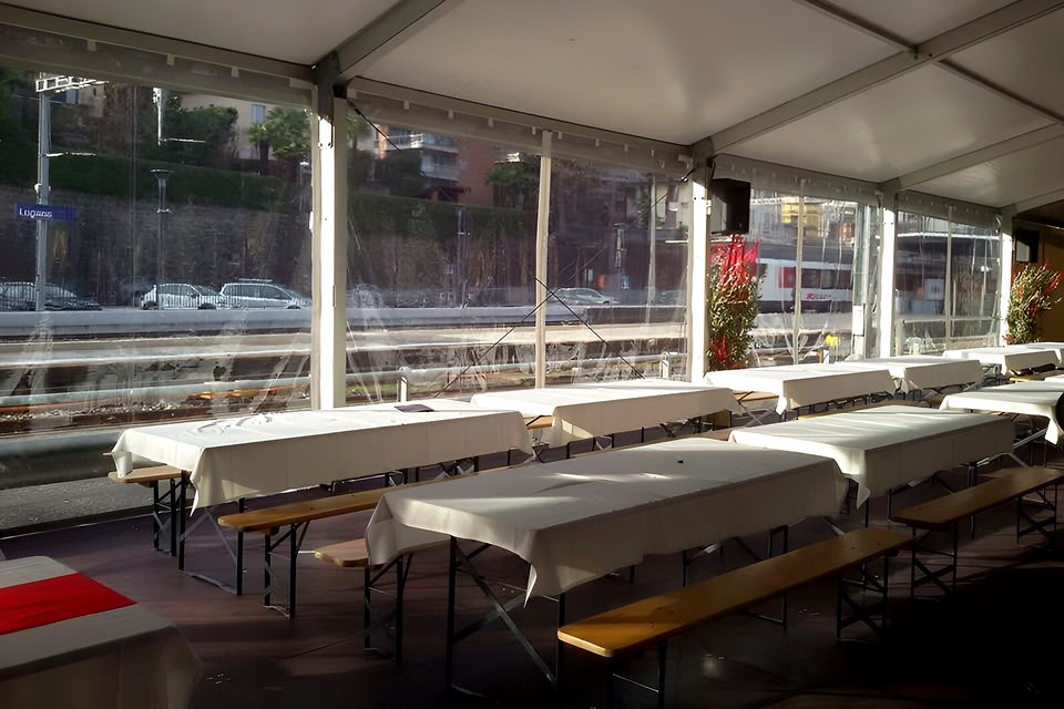 Interno di capannone con tavoli apparecchiati con tovaglie bianche e panche in legno, fuori la stazione di Lugano