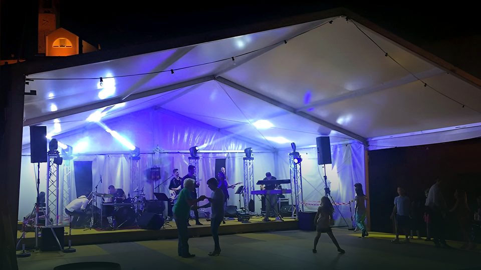 Tensostruttura aperta di notte con una band sul palco, luci blu e viola, gente che balla e un campanile illuminato sullo sfondo