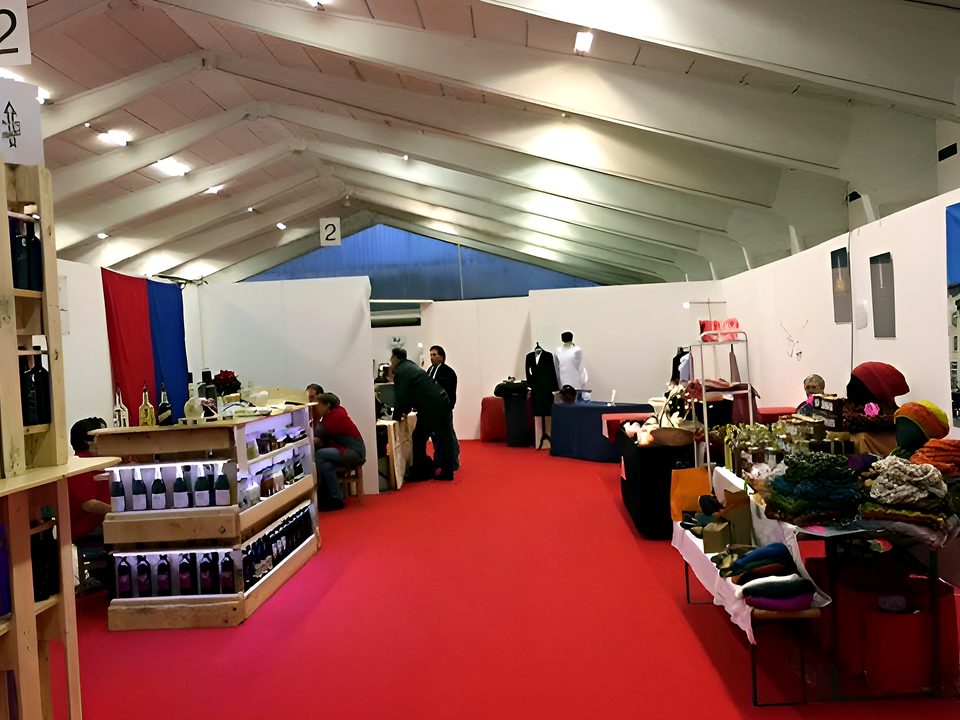 Interno di una grande tensostruttura allestita come fiera, con moquette rossa, stand e visitatori