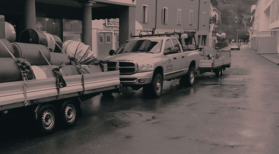 Pick-up con rimorchio carico di fusti di zavorra e teli, fermo davanti a un edificio sotto la pioggia, in bianco e nero
