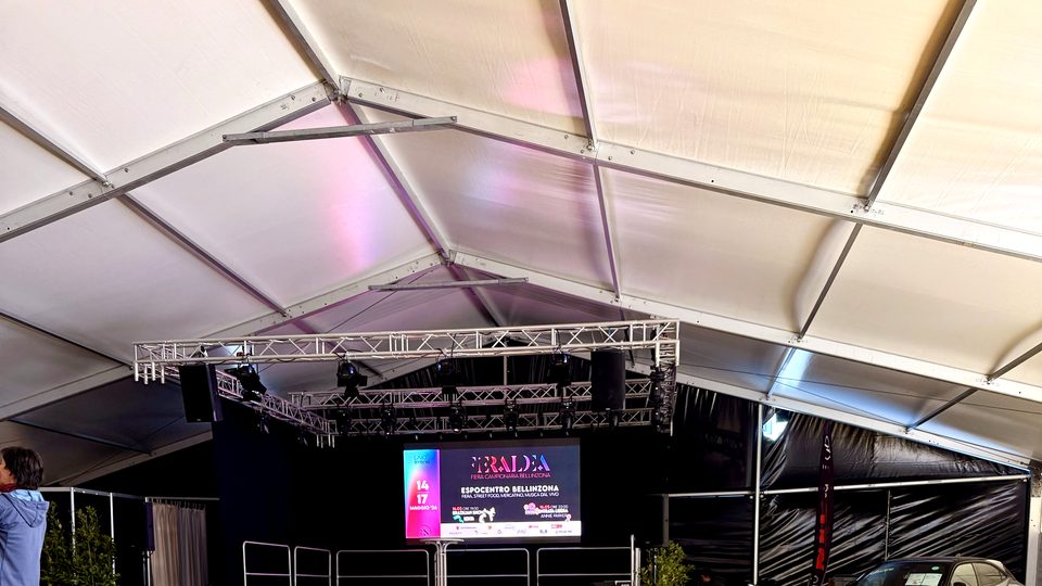 Interno di una grande tensostruttura allestita per una fiera, con schermo led, traliccio luci e capriate in acciaio