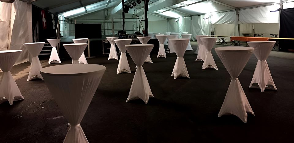 Interno notturno di una tensostruttura con tavoli alti coperti di bianco, palco con traliccio e bandiera svizzera